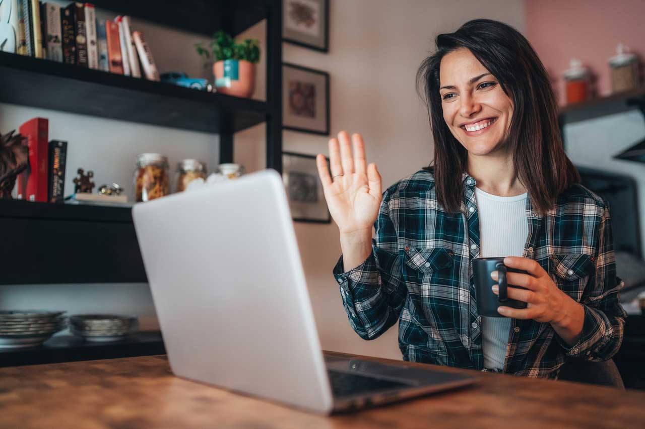 Woman with flannel shirt holding coffee mug waving to laptop near bookshelf