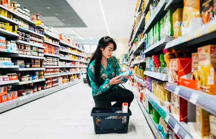 Asian woman kneeling in grocery store reading a label with a basket beneath her. 