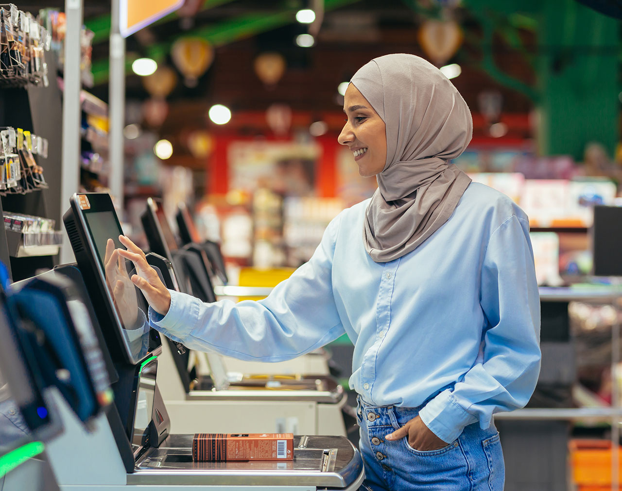 A smiling Muslim woman checks her item out at a self-checkout lane in a store.